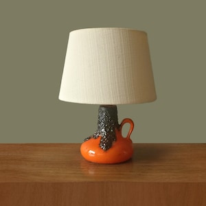 Puede incluir: Una lámpara de mesa con base de cerámica naranja brillante y un esmalte texturizado de color marrón oscuro que gotea desde el cuello. La base incluye una pequeña asa lateral y está rematada con una pantalla de tela color crema.