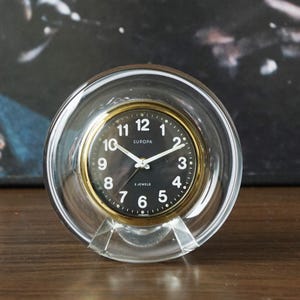 Puede incluir: Un reloj redondo transparente con esfera negra y borde dorado. La esfera muestra números y manecillas blancas, con la palabra "EUROPA" encima del centro. El reloj está sostenido por una base triangular transparente.