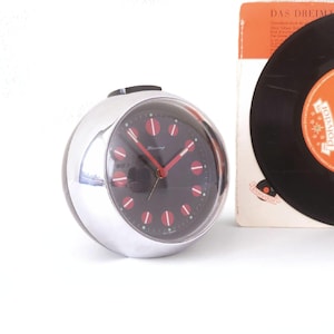 Puede incluir: Un reloj despertador esférico cromado con esfera negra, manecillas rojas e indicadores de hora rojos. La esfera del reloj dice "Blessing". Un disco de vinilo vintage y la portada del álbum son visibles.