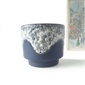 Puede incluir: Una maceta de cerámica azul con un borde texturizado blanco y gris. La maceta tiene forma cilíndrica con una base ligeramente ensanchada. El fondo es blanco, con una imagen enmarcada visible.