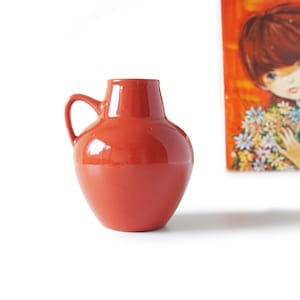 May include: A terracotta-coloured ceramic vase with a handle. The vase has a rounded body and a narrow neck. The glossy finish reflects light. A painting with a girl holding flowers is in the background.
