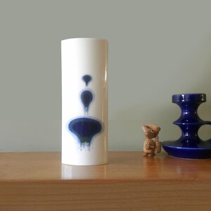 May include: A white ceramic vase with a cylindrical shape. The vase features a blue and dark blue abstract design. A small wooden figurine and a blue candle holder are also visible on a wooden surface.
