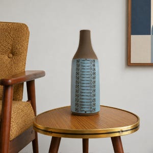 May include: A tall ceramic vase with a smooth brown neck and a cylindrical body featuring a light blue textured pattern of vertical columns and horizontal cross-hatching. The vase has a matte finish and a narrow opening at the top.