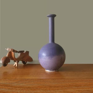 May include: A two-toned purple ceramic vase with a long neck and rounded body. The vase is on a wooden surface, with a wooden scooter figurine to the left. The vase is approximately 10 inches tall.