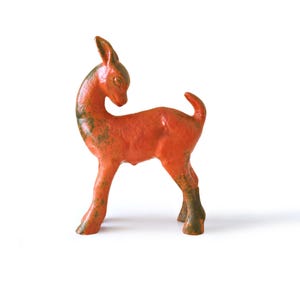 Puede incluir: Figura de ciervo de cerámica en posición de pie, con cuello curvado y cola pequeña. El ciervo es principalmente naranja con acentos verde-marrón en las patas y la espalda. La figura está sobre un fondo blanco.