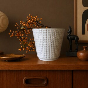Op de afbeelding: Een witte keramische plantenpot met een rasterachtig reliëf aan de buitenkant. De pot is gevuld met een decoratieve tak met talloze kleine, ronde, oranje bessen. De pot heeft een taps toelopende vorm met een gladde, platte onderkant.