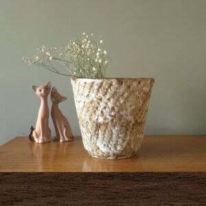Könnte beinhalten: Eine strukturierte, beige Keramikvase mit rauer Oberfläche, die zarte weiße Blumen enthält. Zwei kleine, hellbraune Katzenfiguren sitzen links von der Vase. Die Objekte befinden sich auf einer Holzoberfläche.
