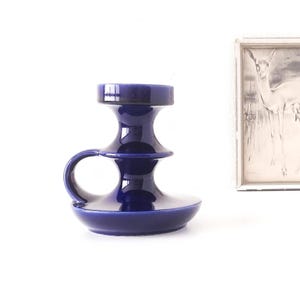 May include: A dark blue ceramic candle holder with a handle. The candle holder features a wide base, a tiered body, and a circular top for a candle. The design is minimalist and modern, suitable for home decor.