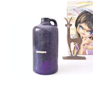 Könnte beinhalten: Eine dunkelviolette Keramikvase mit Henkel. Die Vase hat eine strukturierte Oberfläche mit Craquelé-Effekt und ein Etikett mit der Aufschrift "OZK CERAMIK". Eine Bronze-Hirschfigur und eine Vintage-Illustration befinden sich im Hintergrund.