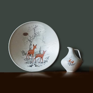 Puede incluir: Un juego de plato y jarra de cerámica blanca. El plato presenta una escena de bosque con dos ciervos, un árbol y follaje en naranja y negro. La jarra tiene un diseño de ciervo similar.