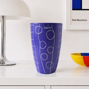 Peut inclure: Un vase cylindrique bleu orné d'un motif de lignes blanches représentant des cercles et des sections en forme de grille verticale. Le mot AMANO est imprimé en blanc près du haut au centre du vase. La surface présente une finition mate.