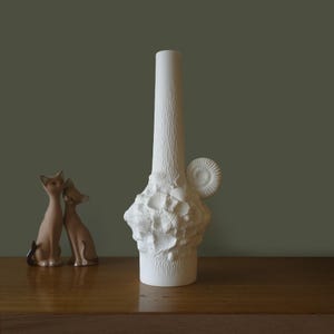 Puede incluir: Un jarrón de cerámica blanca con un cuello alto y cilíndrico y una base texturizada adornada con conchas marinas y diseños en espiral. El jarrón tiene un acabado liso y mate y se encuentra sobre una superficie de madera. Dos figuras de gato están en el fondo.