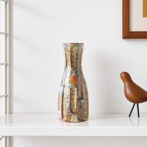 Peut inclure: Un vase en céramique avec un col effilé et un vernis texturé et moucheté dans des tons dorés, bruns, blancs et gris. La surface présente des indentations verticales et une tache rouge orangé, créant un aspect varié et multicolore.