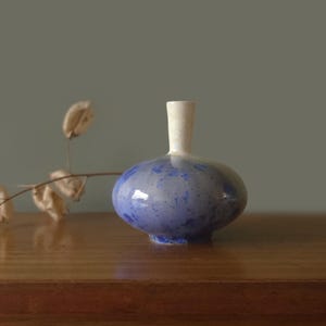 Peut inclure: Un petit vase soliflore en céramique avec un corps arrondi et bulbeux et un col étroit et cylindrique. La base du vase présente un émail tacheté bleu et gris avec des mouchetures bleu foncé, tandis que le col est fini en blanc cassé uni.