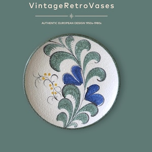 Op de afbeelding: Een rond keramieken bord met een wit gestructureerd oppervlak en een handgeschilderd bloemmotief in groen en blauw, geaccentueerd door kleine gele stippen. De tekst VintageRetroVases en AUTHENTIC EUROPEAN DESIGN 1950s-1980s staat erboven.