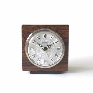 Könnte beinhalten: Eine Vintage Peter Uhr mit silbernem Zifferblatt und römischen Ziffern, eingebettet in einen dunklen Holzquader. Das Zifferblatt hat ein verziertes Blumendesign und schwarze Zeiger. Die Uhr ist ein klassisches Retro-Design.