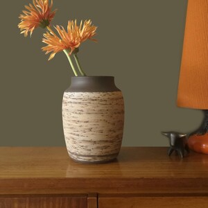 Puede incluir: Un jarrón de cerámica con un exterior texturizado marrón claro y beige y un borde marrón oscuro contiene dos flores naranjas. El jarrón está sobre una superficie de madera, con una lámpara naranja y una pequeña escultura de metal en el fondo.