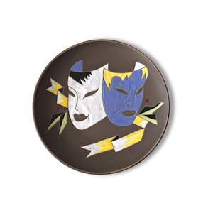 May include: A round dark brown ceramic plate featuring a central design of two theatrical masks, one white and one blue, surrounded by yellow and white ribbon accents with small red dots, creating a bold and colorful graphic composition.