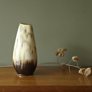 Può includere: Un vaso in ceramica a forma di goccia, con smalto color crema e marrone. Il vaso ha un bordo dorato e presenta gocce blu e rosse. Baccelli secchi poggiano su una superficie di legno.