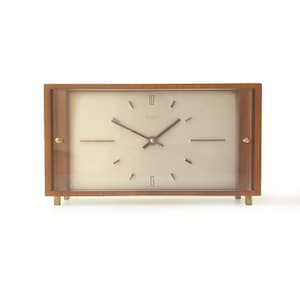 May include: A rectangular mantel clock with a wooden frame and a light-colored face. The clock features thin, dark hands, simple line markers for the hours, and the number 12 and 6 printed on the face. The brand name Kienzle is visible.