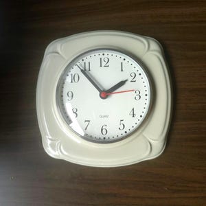May include: A square cream-colored wall clock with rounded corners and a circular face. The clock features black Arabic numerals, black hour and minute hands, a red second hand, and the word QUARTZ printed in black on the white clock face.