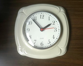 Puede incluir: Un reloj de pared cuadrado de color crema con esquinas redondeadas y una esfera circular. El reloj presenta números arábigos negros, manecillas de hora y minutos negras, un segundero rojo y la palabra QUARTZ impresa en negro en la esfera.