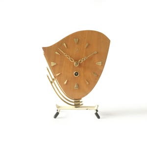 Könnte beinhalten: Eine Vintage-Holzuhr mit hellbraunem Zifferblatt und goldfarbenen Zeigern und Markierungen. Die Uhr hat eine einzigartige, geschwungene Form und steht auf einem weißen Sockel mit goldenen und schwarzen Akzenten. Das Zifferblatt hat ein kleines, kreisförmiges schwarzes Detail.
