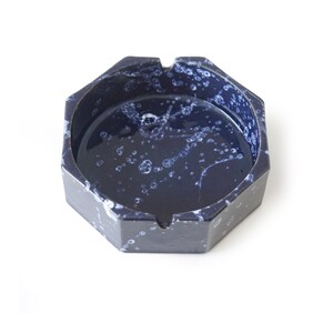 Op de afbeelding: Een marineblauwe keramische asbak met een achthoekige vorm. De asbak heeft een gespikkeld wit en blauw patroon aan de binnen- en buitenkant. Het heeft drie groeven voor het plaatsen van sigaretten.