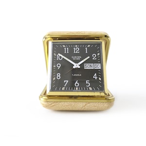 May include: A vintage Europa Day Date travel alarm clock with a gold-tone case and a black face. The clock displays the time, day, and date. The clock is approximately 3 inches tall and 3 inches wide.