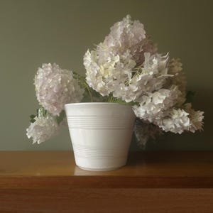 Op de afbeelding: Een witte keramische plantenpot met horizontale ribbeldetails aan de buitenkant, gevuld met een grote, overvolle bos lichtroze en witte hortensiabloemen met groene stelen die zichtbaar zijn in de pot.