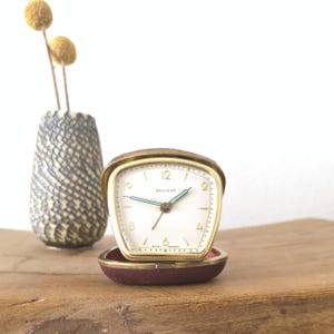 Puede incluir: Un reloj despertador de viaje vintage dorado con esfera blanca y manecillas verdes, marcado "BALVEET" y "WEST GERMANY". El reloj está abierto, revelando una caja burdeos. Un jarrón decorativo con flores amarillas está en el fondo.