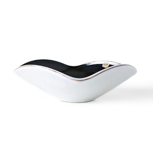 May include: A white ceramic bowl with a black interior and a wavy, asymmetrical shape. The rim has a thin, brown accent. The bowl is designed with a modern aesthetic and is suitable for serving or display.