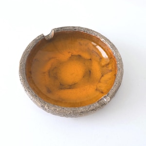 Puede incluir: Un cenicero redondo de cerámica naranja con una textura rugosa similar a la corteza de un árbol alrededor del borde.