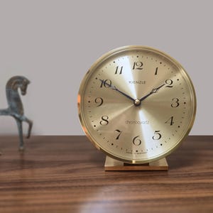 Puede incluir: Un reloj Kienzle chronoquartz dorado con números y manecillas negras. El reloj tiene una esfera redonda y se apoya sobre una base rectangular. Las palabras "chronoquartz" y "Made in Germany" son visibles.