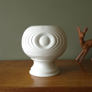 May include: A white ceramic vase with an eye-shaped design. The vase has a round body with concentric, wave-like patterns and a pedestal base. A small, brown deer figurine is in the background.