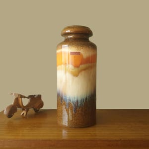 May include: A tall cylindrical ceramic vase featuring a gradient glaze that transitions from a dark brown base to a creamy white middle, followed by a vibrant orange band near the top. A small rectangular label with the text Scheurich is visible.