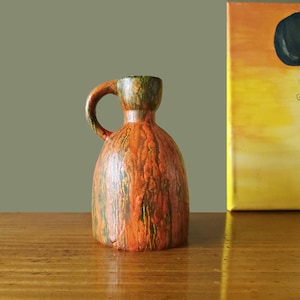 Può includere: Piccolo brocca decorativa in ceramica con manico. Presenta colori arancioni con striature marroni, verdi e gialle. Posizionata su una superficie in legno, con un dipinto sullo sfondo.