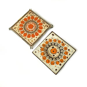 Puede incluir: Dos salvamanteles cuadrados de cerámica con diseños florales en naranja y marrón. Los salvamanteles tienen un fondo blanco con un patrón repetido de círculos naranjas y un centro marrón. Uno de los salvamanteles tiene un marco de metal.