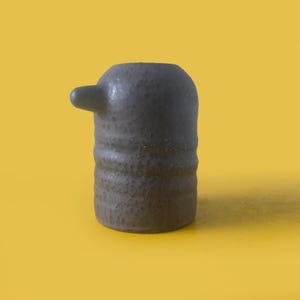 May include: A cylindrical ceramic vessel in a dark charcoal gray hue with a textured, speckled surface. The object features a single protruding spout on the upper left side, creating a distinct asymmetrical silhouette with a flat base.