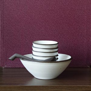 May include: A white ceramic bowl with a dark rim and a stack of smaller bowls. A dark-handled spoon rests in the bowl. The bowls are on a dark wooden surface against a burgundy background.