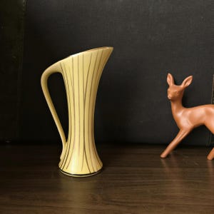 May include: A small, yellow ceramic vase with gold stripes and a curved handle. The vase is sitting on a wooden surface.