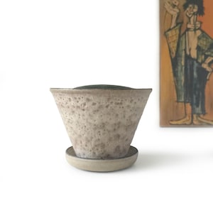 Op de afbeelding: Een gespikkelde, beige keramische plantenbak met een uitlopende vorm, die op een bijpassende schotel staat. De binnenkant van de plantenbak is donkergroen. De plantenbak heeft een textuur en een rustieke uitstraling. Een schilderij staat op de achtergrond.