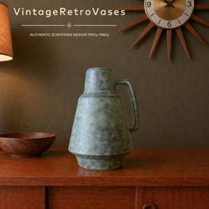 May include: A ceramic pitcher vase with a textured, mottled green and grey finish. The vessel features a wide, tapered base, a narrow neck, and a single angular handle on the right side. The text VintageRetroVases and AUTHENTIC EUROPEAN DESIGN 1950s-1980s is visible.