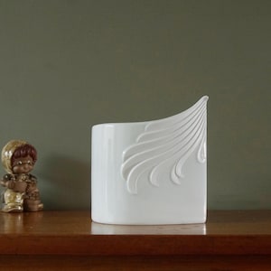 May include: A white ceramic vase with a curved, wing-like design on one side. The vase has a rectangular shape and sits on a wooden surface. A small figurine is to the left of the vase.