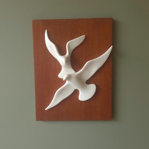 May include: A rectangular wall plaque featuring two white ceramic birds in flight mounted on a smooth, dark brown wooden board. The birds are sculpted with outstretched wings, creating a three-dimensional effect against the flat, solid-colored wood background.