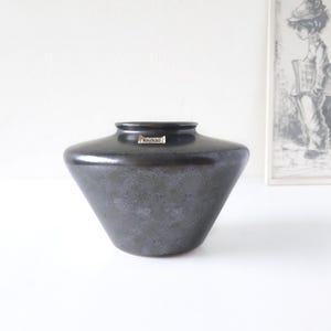 Puede incluir: Un jarrón de cerámica negro con una boca ancha y una base acampanada. El jarrón tiene un esmalte moteado y una pequeña etiqueta que dice "MOXA".