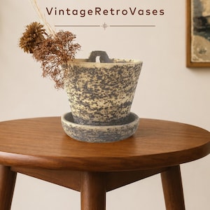Peut inclure: Un pot en céramique avec une finition mouchetée grise et crème, doté d'une petite anse sur le bord supérieur et d'une soucoupe circulaire assortie. Le pot contient des tiges florales brunes séchées et affiche le texte VintageRetroVases.