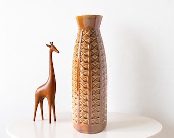 May include: A tall, cylindrical ceramic vase with a glossy brown glaze and a repeating geometric diamond pattern across its surface, positioned next to a smooth, carved wooden giraffe figurine with a long neck and slender legs.