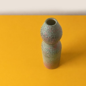 May include: A ceramic vase with a speckled green and pink finish. The vase features a distinct hourglass shape with a rounded upper section and a cylindrical base, showcasing a textured, mottled surface pattern throughout the entire exterior.