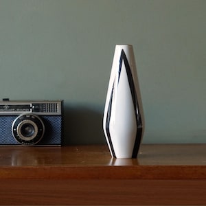 Peut inclure: Un vase en céramique blanc avec un motif géométrique noir. Le vase a une forme haute et élancée avec une ouverture étroite. Un appareil photo vintage est à gauche du vase sur une surface en bois.